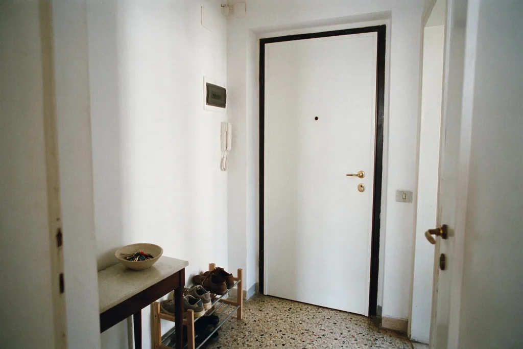 Un ingresso di appartamento con porta bianca, un mobile con scarpe e una ciotola con le chiavi, pavimento in graniglia
