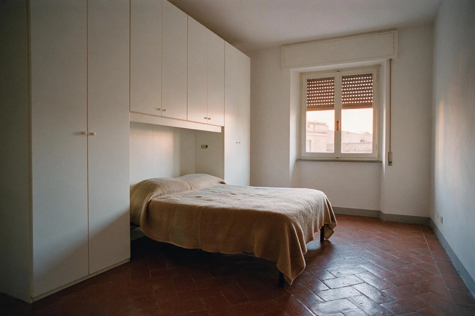 Una camera da letto con armadio a ponte bianco sopra il letto, pavimento in cotto a spina di pesce e finestra con tapparelle