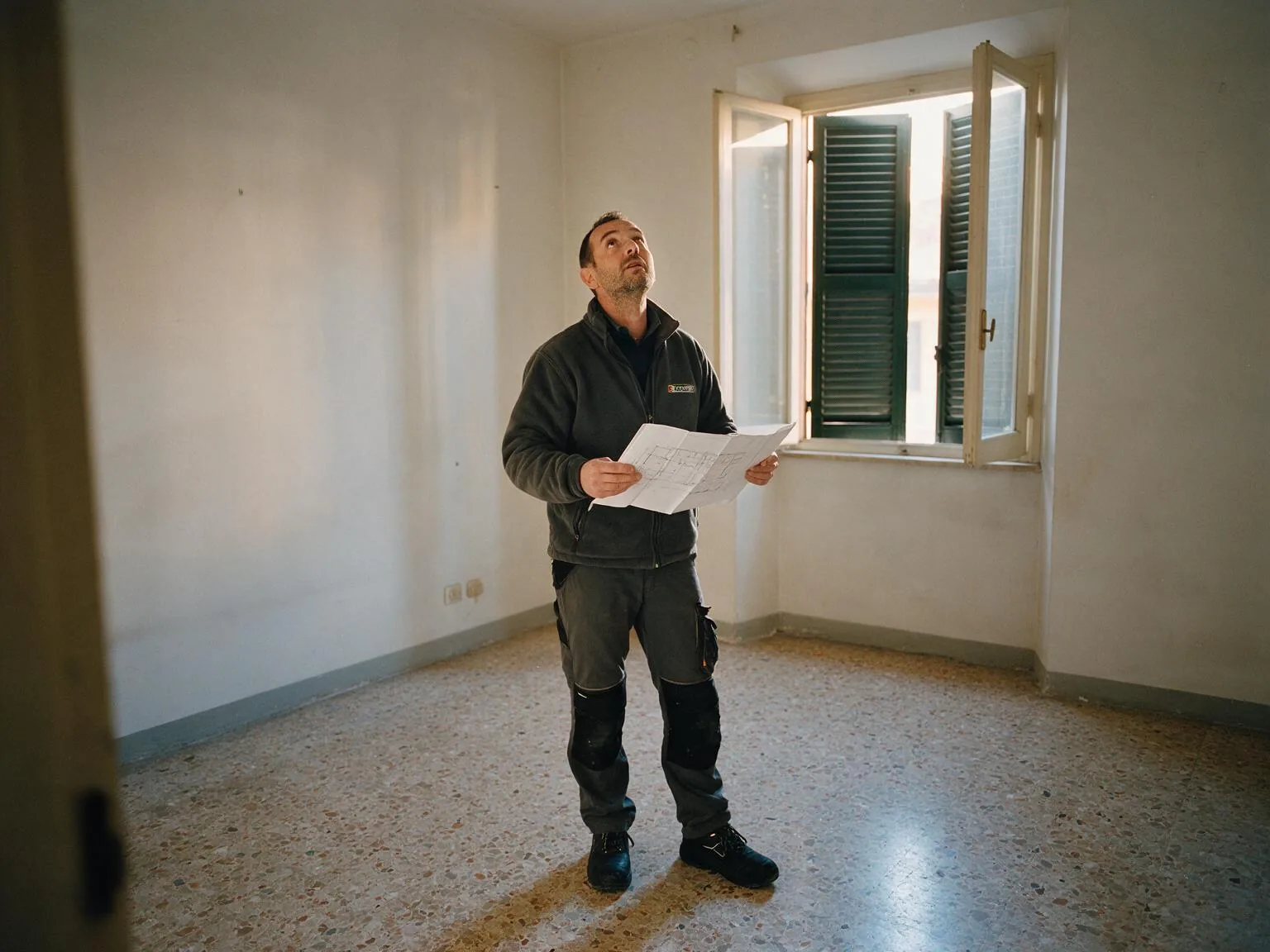 Un tecnico in una stanza vuota con la pianta dell'appartamento in mano guarda verso il soffitto