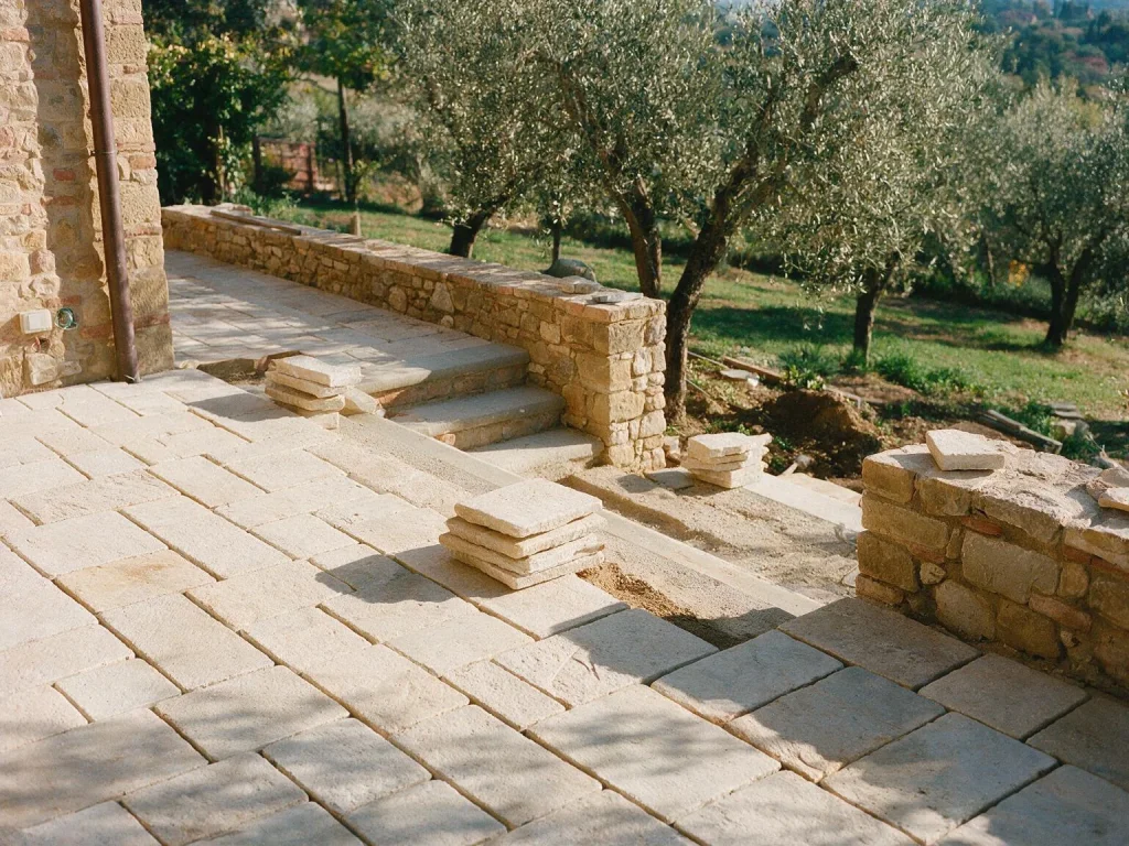 Una terrazza di villa con il nuovo pavimento in pietra in posa e una scala che scende verso un uliveto.